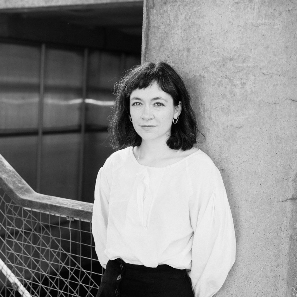 a black and white image of a person wearing a white top and dark trousers. shoulder-length hair and a fringe leaning against a wall